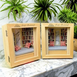Op de afbeelding: Twee houten schaduwdozen met glazen fronten, die elk een miniatuur boekenkast met een leeshoek tonen. De dozen zijn gelabeld "Judy's Bookshelf" en bevatten een boek met een bloemenontwerp.
