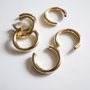 May include: Several gold-colored hoop earrings are scattered on a white surface. Some are open, while others are interlocked. The earrings have a shiny, metallic finish, reflecting the light.