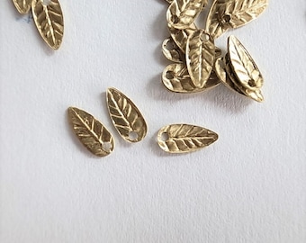 May include: A pile of gold-colored metal leaf charms with a hole in the center of each leaf. The leaves have a textured surface and are shaped like a teardrop.
