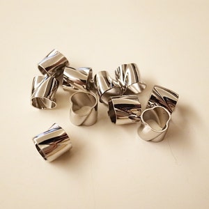 Könnte beinhalten: Eine Sammlung glänzender, silberfarbener Serviettenringe. Jeder Ring hat ein einzigartiges, verdrehtes Design, das einen modernen und eleganten Look erzeugt. Die Ringe sind auf einer hellen Oberfläche verstreut, was ihre reflektierenden Eigenschaften hervorhebt. Ideal für Tischdekorationen.