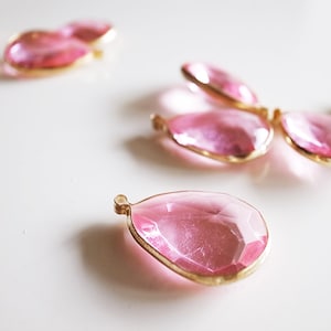 Könnte beinhalten: Nahaufnahme mehrerer rosafarbener, tropfenförmiger Anhänger mit goldfarbenen Metallrändern. Die Anhänger sind facettiert und fangen das Licht ein. Der Hintergrund ist eine schlichte, weiße Oberfläche. Diese Anhänger eignen sich für die Schmuckherstellung.