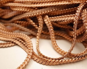 May include: A close-up of a pile of copper-colored chain links. The chain is made of small, flat, interlocking links that are twisted together to create a rope-like effect.