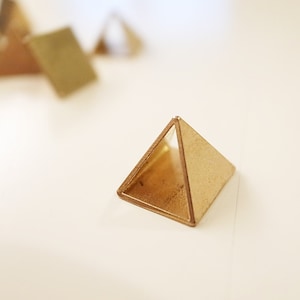 Puede incluir: Una pirámide de metal de color dorado con un centro hueco. La pirámide está sobre una superficie blanca.