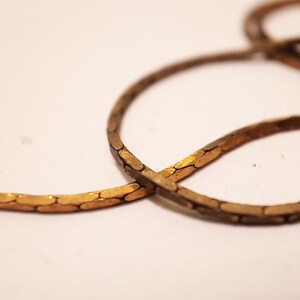 Op de afbeelding: Close-up van een goudkleurige ketting met een plat, in elkaar grijpend ontwerp. De ketting is licht gebogen en ligt tegen een witte achtergrond, wat de metaalglans en het ingewikkelde patroon laat zien.