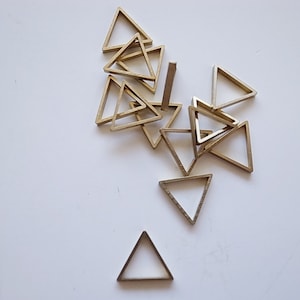 Könnte beinhalten: Eine Ansammlung kleiner, goldfarbener Metall-Dreiecke, die auf einer weißen Oberfläche verstreut sind. Die Dreiecke sind offen gerahmt und haben ein einfaches, geometrisches Design. Die Formen sind wahrscheinlich Komponenten für Schmuck- oder Bastelprojekte.