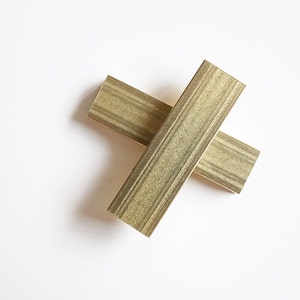 May include: Two gold-coloured rectangular wooden pieces are arranged in a crossed position against a white background. Each piece has a series of parallel, darker lines running lengthwise, creating a textured appearance. The wood has a slightly weathered look.