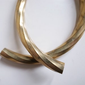 May include: A pair of gold-colored, twisted metal earrings. The earrings have a textured, ridged design and a polished finish. The ends of the earrings are flat and angular. The background is white.