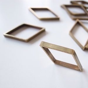 May include: Several small, open, brass-colored diamond-shaped metal frames are scattered on a white surface. The frames are made of flat, rectangular bars, and are slightly angled, creating depth and dimension.