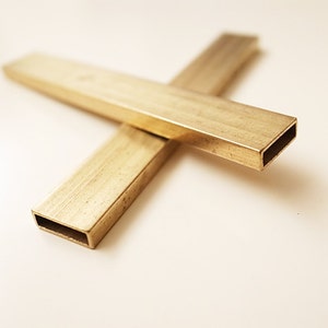 Puede incluir: Dos barras de metal de color dorado con secciones transversales cuadradas están dispuestas en forma de cruz. Las barras son lisas y tienen un acabado ligeramente brillante.