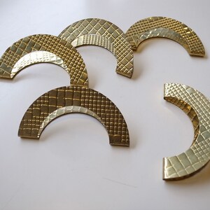 May include: A collection of five brass-toned, semi-circular cabinet pulls. Each pull features a textured surface with a repeating pattern, and a smooth, curved inner edge. The pulls are arranged on a white surface, showcasing their decorative design.