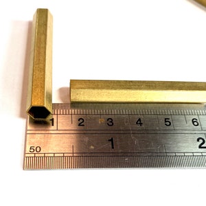 May include: Two brass hexagonal rods, one measuring 15 cm long and the other measuring 2.5 cm long, are placed on a ruler.