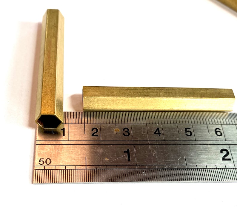 May include: Two brass hexagonal rods, one measuring 15 cm long and the other measuring 2.5 cm long, are placed on a ruler.