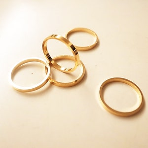 Puede incluir: Una colección de anillos dorados, cada uno con un diseño de banda simple y plana. Los anillos están dispersos sobre una superficie de color claro, con diferentes ángulos y sombras que resaltan su brillo metálico. Los anillos parecen ser de tamaño similar.