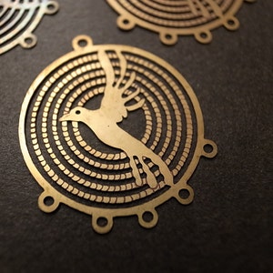 May include: A brass pendant with a circular design featuring a stylized bird in flight. The bird is surrounded by concentric circles, creating a decorative and intricate pattern.
