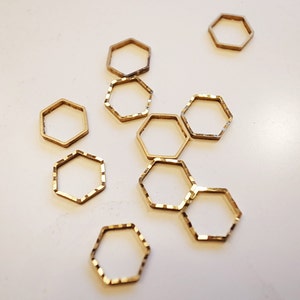 May include: A collection of small, gold-colored, hexagonal metal rings scattered on a white surface. Some rings have a smooth finish, while others have a textured, faceted design. The rings are likely jewelry components or craft supplies.