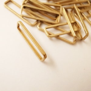 May include: A pile of gold-colored metal rectangular rings, some are stacked on top of each other, and one ring is laying flat on a white surface.