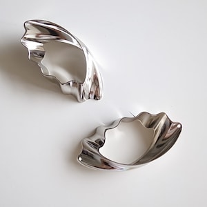 May include: Two silver-colored, abstract-shaped earrings. Each earring has a wavy, curved design with a polished, reflective surface. The earrings are set against a white background, highlighting their form and shine.