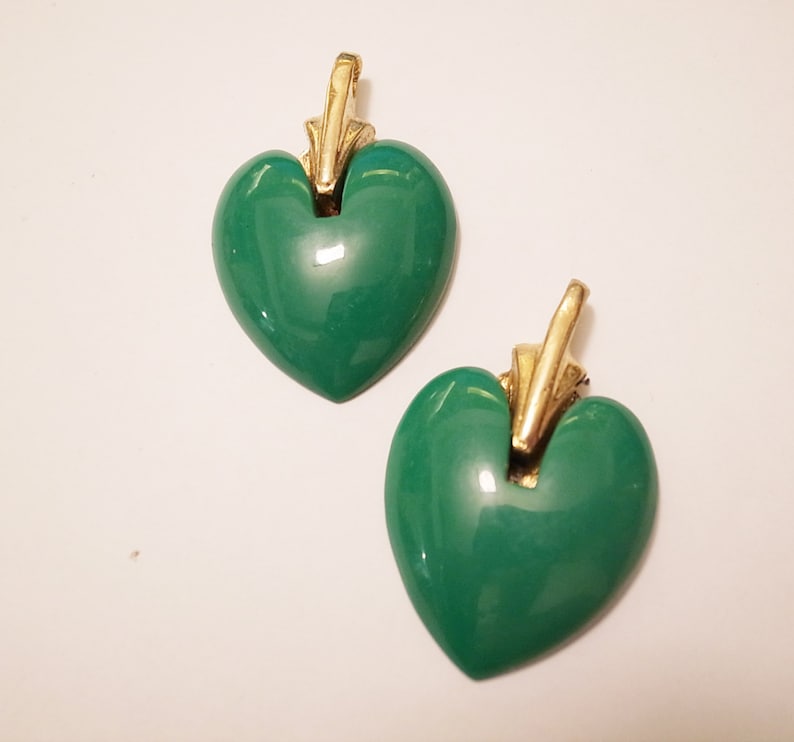 Puede incluir: Un par de pendientes colgantes en forma de coraz&oacute;n verde con detalles dorados. Los pendientes tienen un acabado brillante y lustroso.