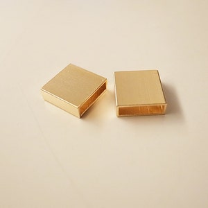 Könnte beinhalten: Zwei quadratische Verbinder aus goldfarbenem Metall mit einer rechteckigen Öffnung in der Mitte.