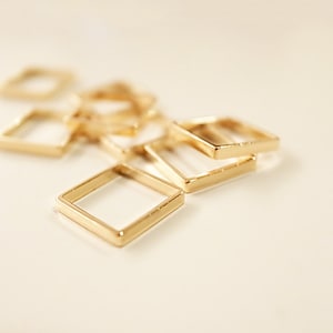 Puede incluir: Primer plano de varios pequeños marcos cuadrados dorados. Los marcos están dispersos sobre una superficie clara, algunos enfocados y otros borrosos en el fondo. Los marcos parecen ser de metal y tienen un acabado brillante.