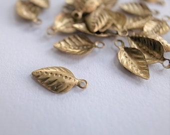 May include: Close-up of several small, gold-colored leaf charms. Each charm has detailed veining and a small loop at the top for attaching to jewelry or other crafts. The leaves are scattered on a white surface.