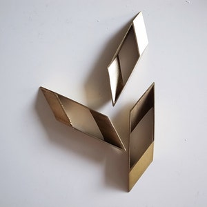 Op de afbeelding: Drie geometrische objecten van goudkleurig metaal, gerangschikt op een wit oppervlak. Elk object heeft een ruitvorm met een holle binnenkant en een contrasterende zilverkleurige streep. De objecten zijn schuin geplaatst.