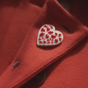 Op de afbeelding: Een rode wollen jas met een witte hartvormige broche. De broche heeft een ontwerp van kleine rode hartjes.
