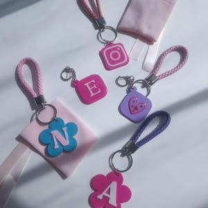 Pode incluir: Uma coleção de chaveiros com alças trançadas e argolas de metal. Os chaveiros apresentam várias formas, incluindo uma flor, um coração e o logotipo do Instagram. As cores incluem rosa, roxo e azul.