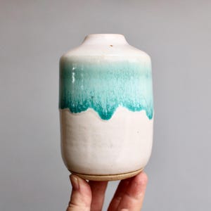 May include: A handmade ceramic vase with a white base and a turquoise, dripped glaze. The vase is cylindrical with a rounded top and measures approximately 5 inches tall.