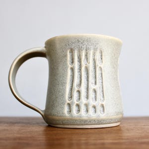 May include: A speckled, stoneware mug with a light gray glaze. The mug features a curved handle and a vertical, textured design on the front. The mug is sitting on a wooden surface.