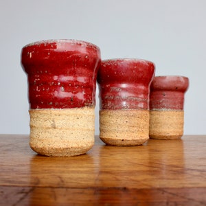 May include: Three red and cream ceramic mugs with a textured glaze. The mugs are arranged in a row on a wooden surface.