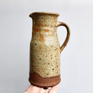 May include: A speckled brown and beige ceramic pitcher with a wide mouth and a handle. The pitcher has a rustic, handmade look.