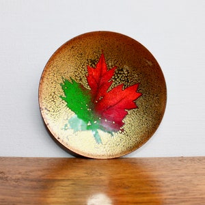 May include: A small, round, copper plate with a red and green maple leaf design. The plate has a gold background with a textured finish.