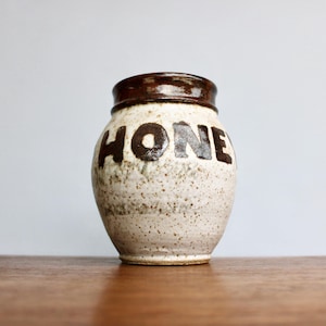 May include: A speckled, off-white ceramic honey pot with a dark brown rim and the word "HONEY" in dark brown block letters. The jar has a rustic, handmade appearance and sits on a wooden surface.