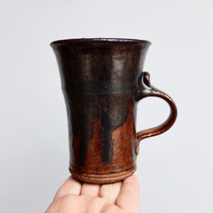 May include: A brown and black ceramic mug with a flared rim and a handle. The mug has a rustic, handmade look.