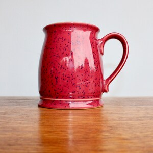 May include: A red speckled ceramic mug with a wide base and a rounded handle.