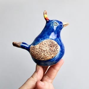 May include: A ceramic bird figurine with a glossy blue finish. The bird has a textured brown patch on its chest and tail, a yellow, red, and blue crest, and a light blue eye. The beak is a light tan color.