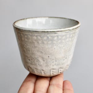 May include: A handmade ceramic cup with a textured, off-white exterior and a smooth, white interior. The cup features a slightly flared shape and a decorative band near the top. The word "PEACE" is embossed on the bottom.