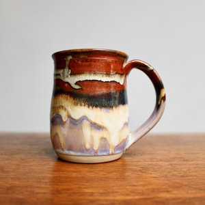May include: A handmade ceramic mug with a unique glaze. The mug features a blend of colors, including brown, white, and purple, with a glossy finish. The mug has a large handle and is suitable for hot or cold beverages.