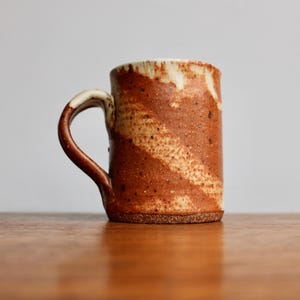 May include: A brown and white ceramic mug with a speckled glaze. The mug has a rounded body and a handle.