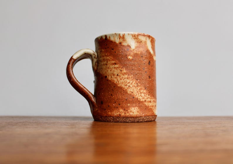 May include: A brown and white ceramic mug with a speckled glaze. The mug has a rounded body and a handle.