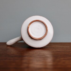 May include: White ceramic pitcher with a brown rim and a handle. The pitcher is sitting on a wooden surface.