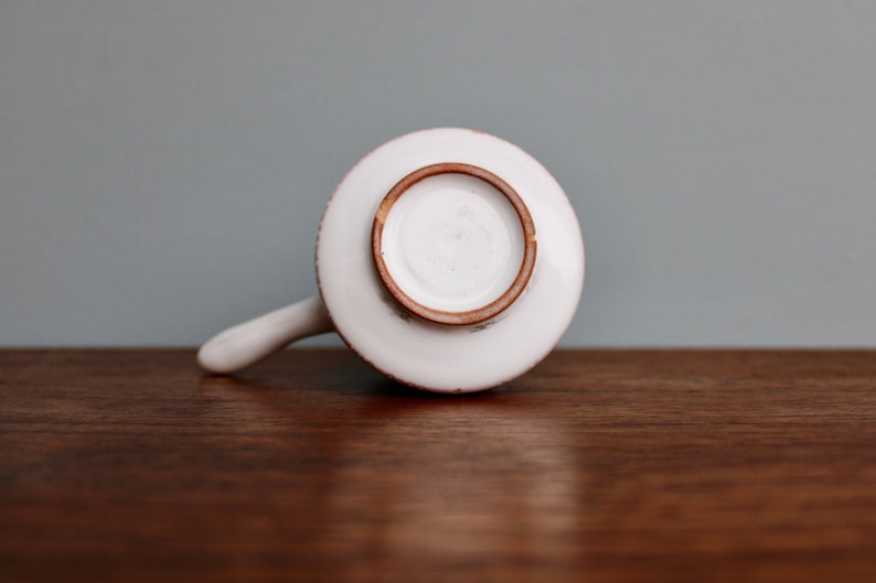 May include: White ceramic pitcher with a brown rim and a handle. The pitcher is sitting on a wooden surface.