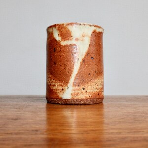 May include: A brown ceramic mug with white glaze, featuring a unique abstract design. The mug has a speckled texture and a slightly rounded shape.