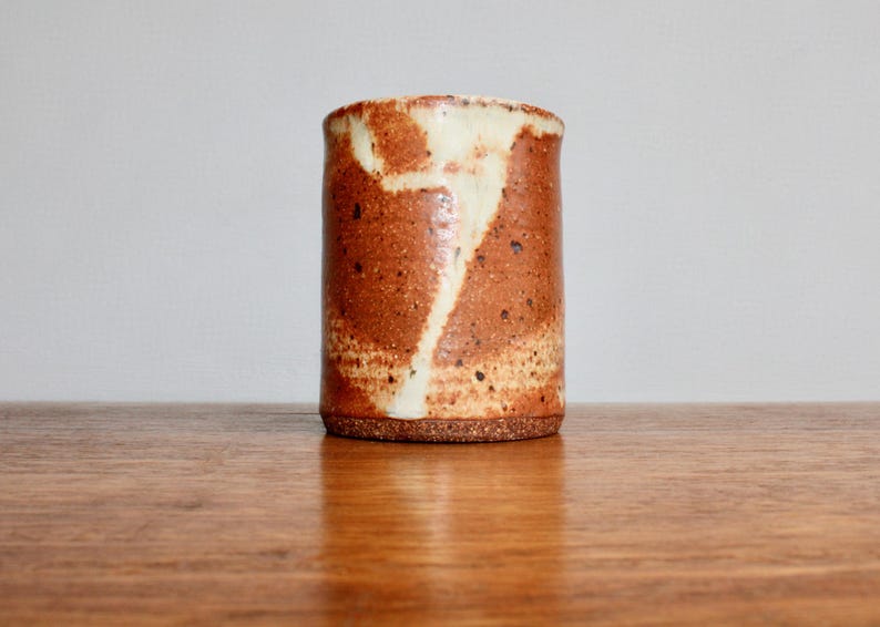 May include: A brown ceramic mug with white glaze, featuring a unique abstract design. The mug has a speckled texture and a slightly rounded shape.