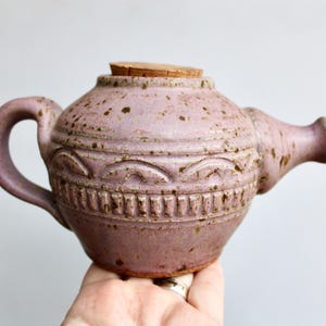 May include: A handcrafted, lavender-colored ceramic teapot with a cork stopper. The teapot features a rounded body with a decorative band of carved patterns and a curved handle. The spout is short and stubby. The surface has a speckled texture.