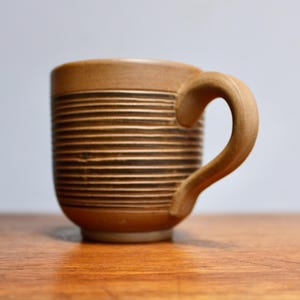 May include: A brown ceramic mug with horizontal black stripes and a curved handle. The mug is on a wooden surface. The mug is a light brown color with a darker brown stripe design.