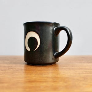 Pu&ograve; includere: Una tazza in ceramica nera con un design a forma di luna crescente bianca. La tazza ha un manico arrotondato.