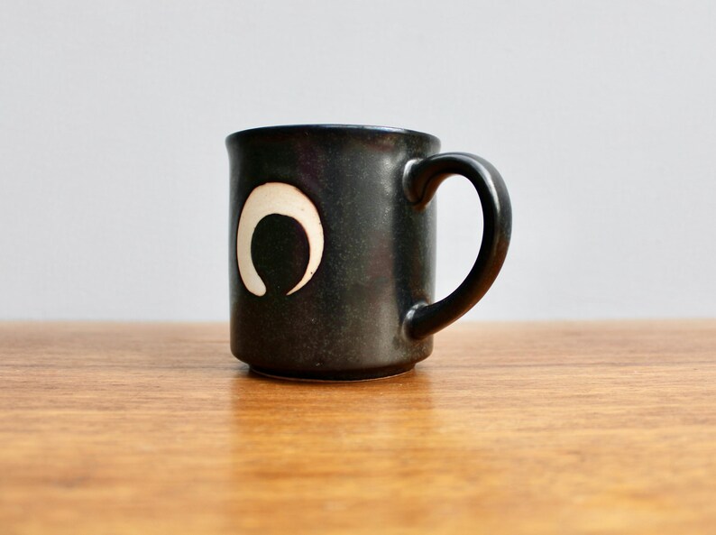 Pu&ograve; includere: Una tazza in ceramica nera con un design a forma di luna crescente bianca. La tazza ha un manico arrotondato.
