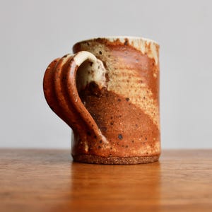 May include: A brown and white ceramic mug with a unique, twisted handle. The mug has a speckled glaze and a rustic look.